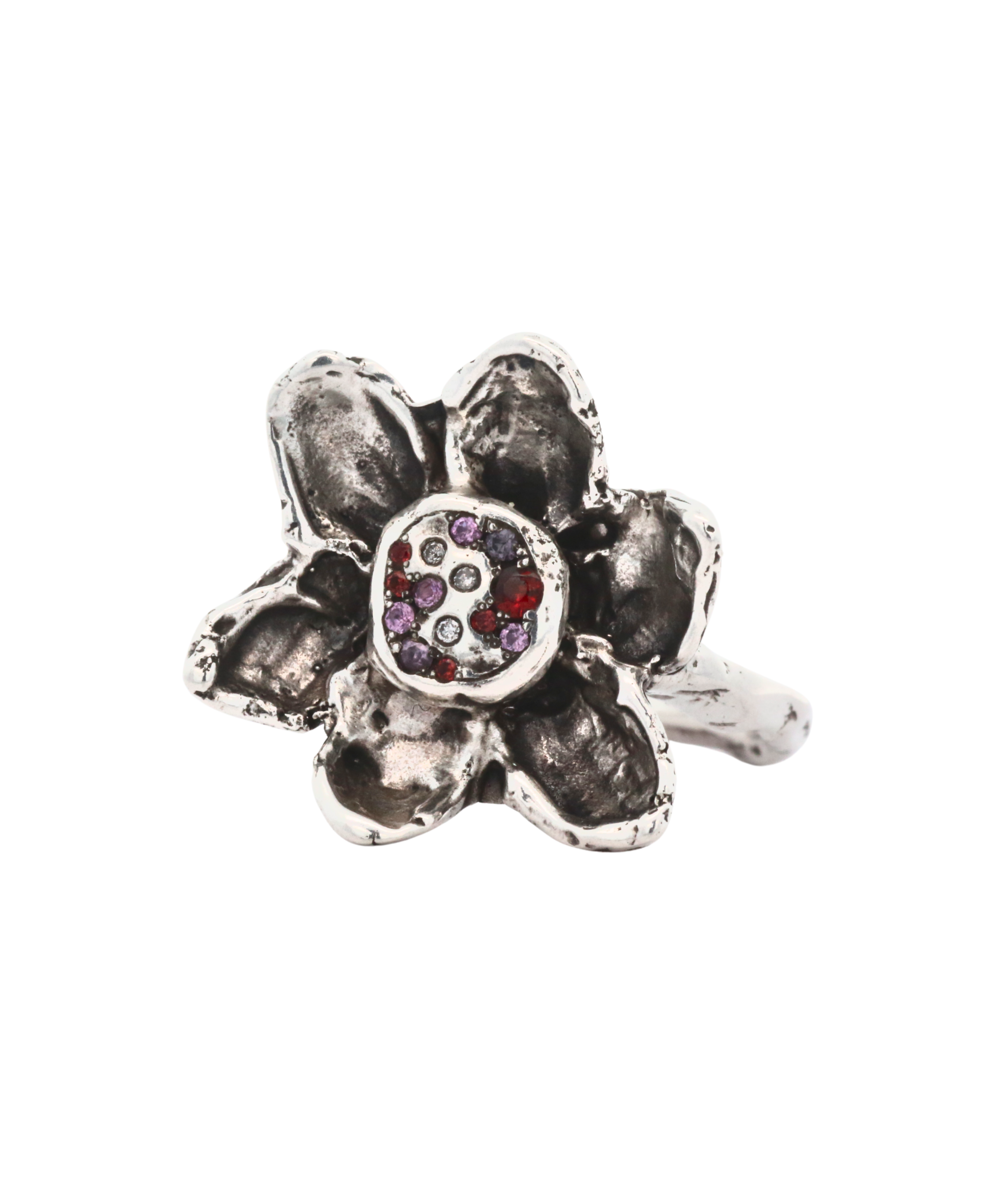 A silver-toned flower ring with textured petals and a central cluster of small gemstones in red, purple, and clear, shown on a plain white background.